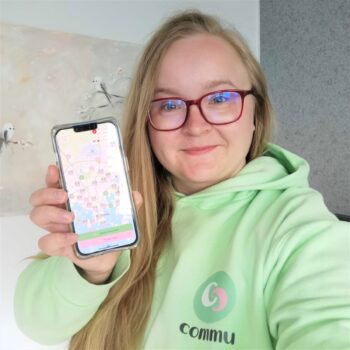 Karoliina Kauhanen, founder of Commu, smiles in a green hoodie while holding up a smartphone displaying the Commu app.