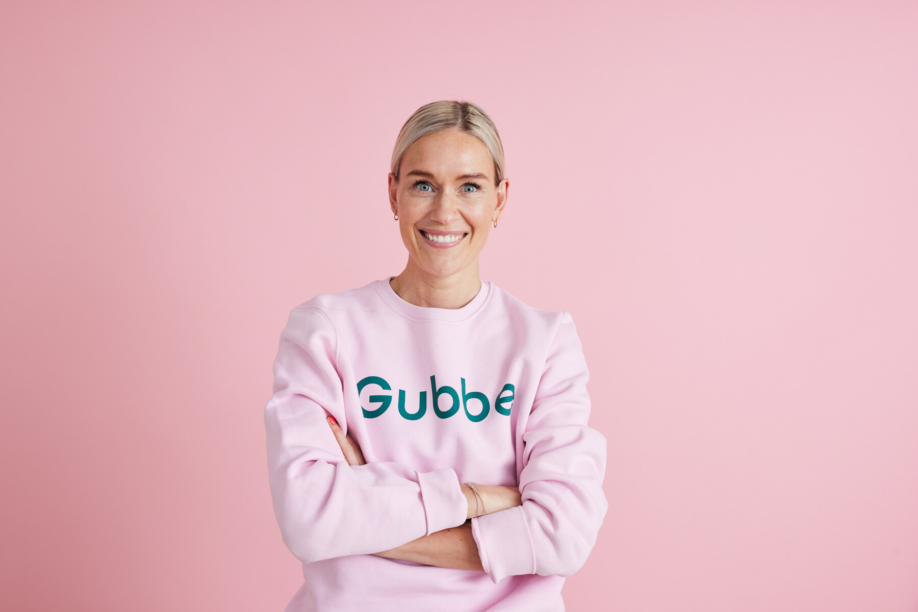 A woman, Meri-Tuuli Helin, CEO of Gubbe, stands smiling with her arms crossed against a pink background, wearing a light pink Gubbe sweatshirt.