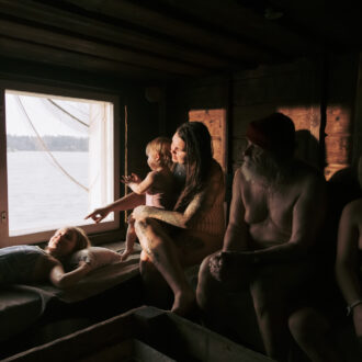 Varias personas de distintas edades, con trajes de baño o aparentemente sin ropa, están sentadas en el interior en penumbra de una sauna de paredes de madera, con ventanas desde las que puede verse un lago.