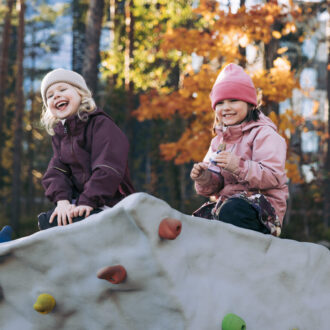 Happiness in Finland: Two young, smiling children in warm clothing are sitting atop a large rock on a playground with autumn foliage showing in the background.