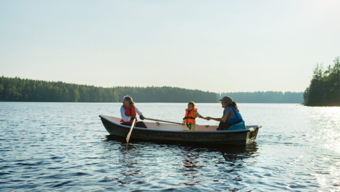 Happiness in Finland: Two adults and a small child, all in lifejackets, are crossing a calm, forest-lined lake in a rowboat.