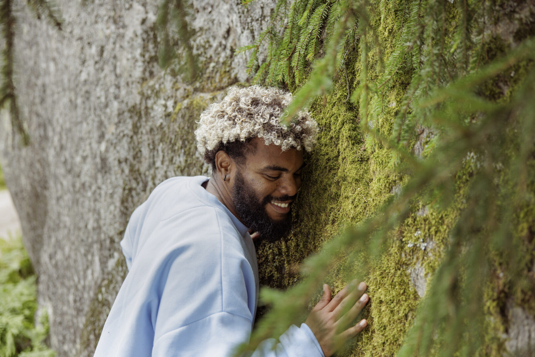 A smiling man is leaning his cheek, chest and hand against a vertical rock surface covered with moss in a forest.