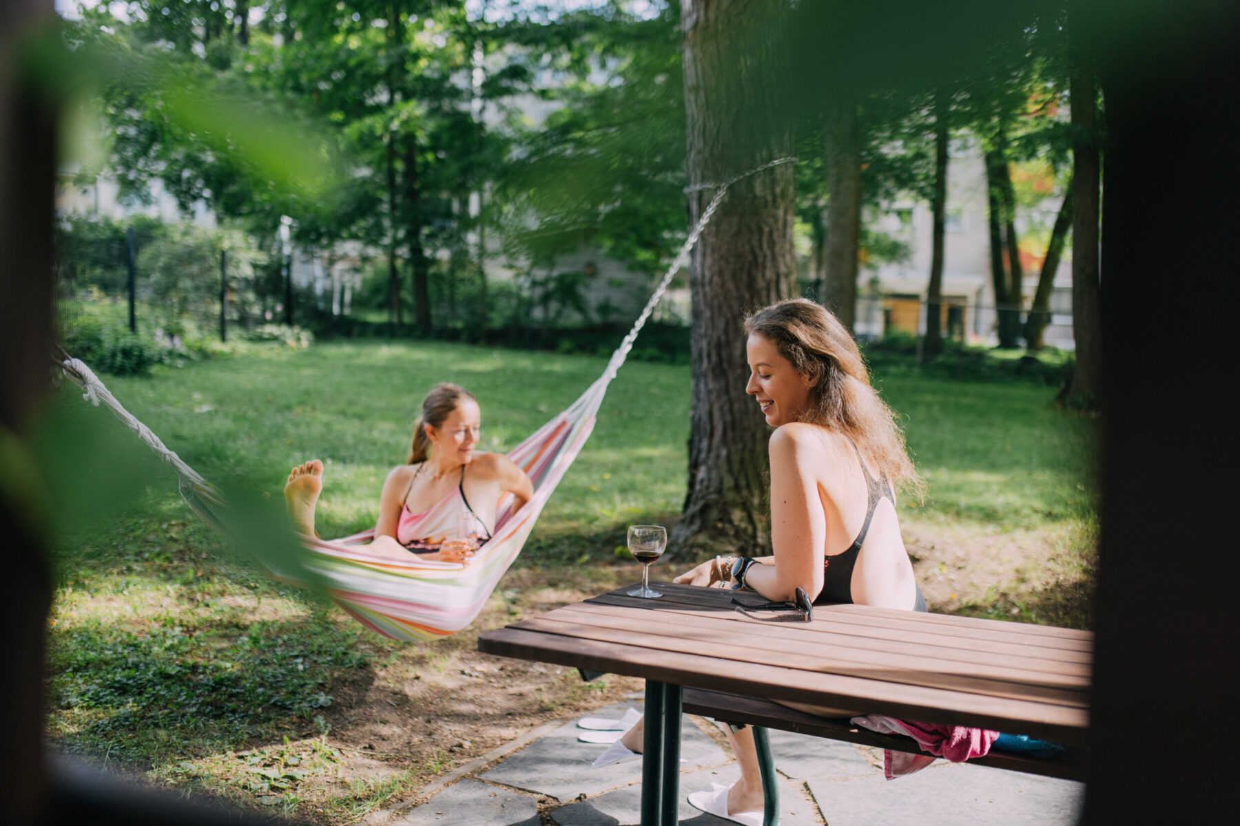 Two women in bathing suits, one lying in a hammock and one sitting at a table, relax in the grassy yard of an apartment building on a sunny day.