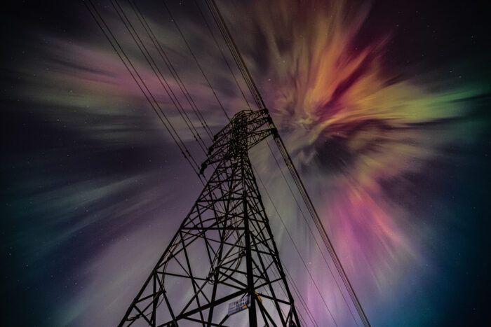 In a view from the base of a power transmission tower, vibrant aurora borealis spreads across the night sky in streaks of green, pink and gold.