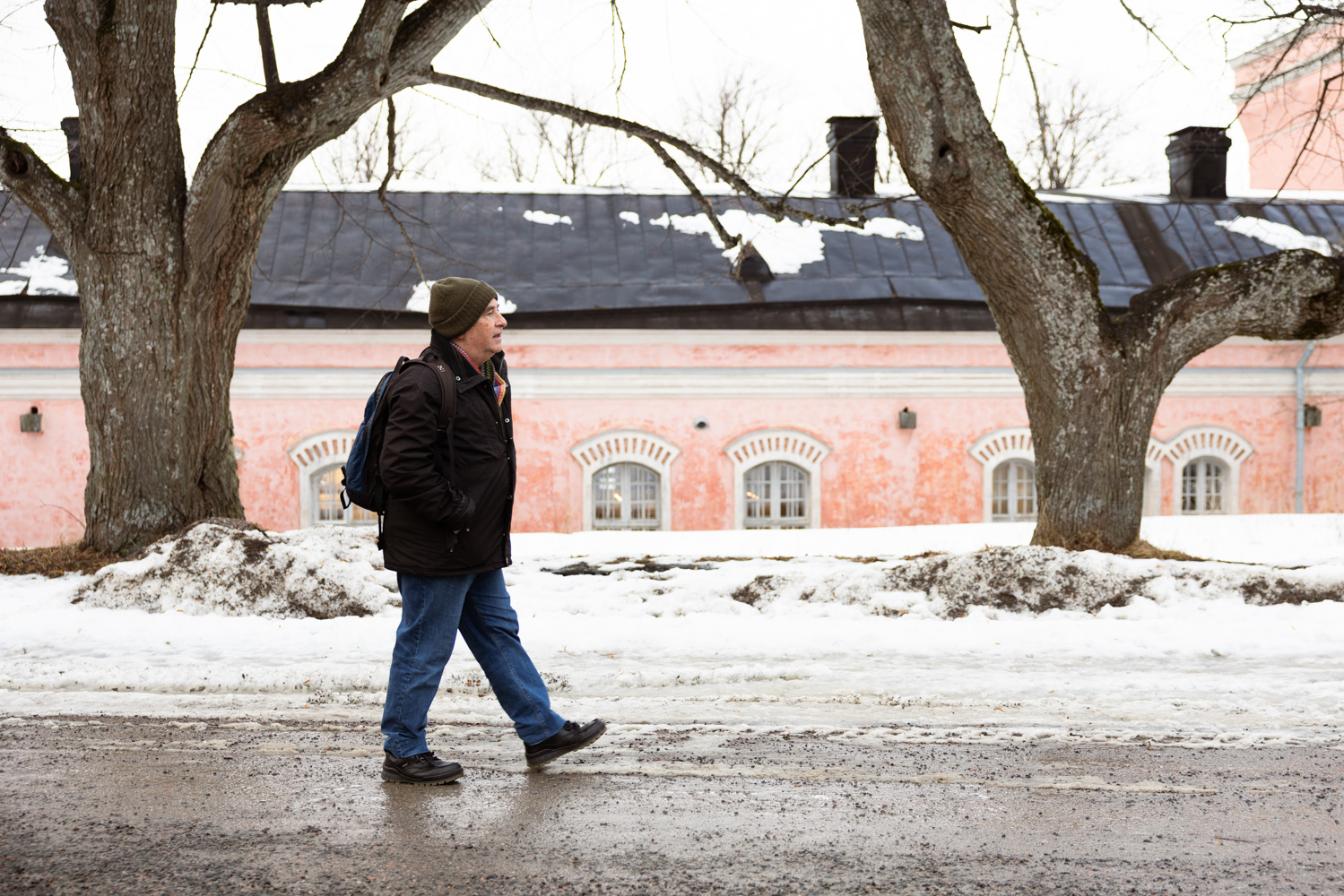 Tim Bird walks between bare trees with snow on the ground in front of a pink building at Suomenlinna.