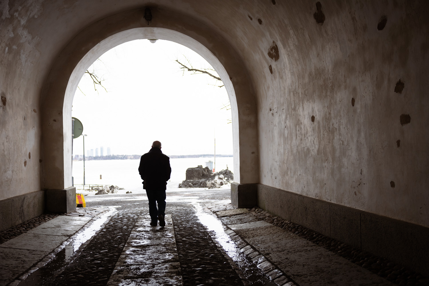 Tim Bird, seen from behind, walks along a cobbled path through an arched tunnel on Suomenlinna, with snow, the frozen sea and distant city buildings ahead.