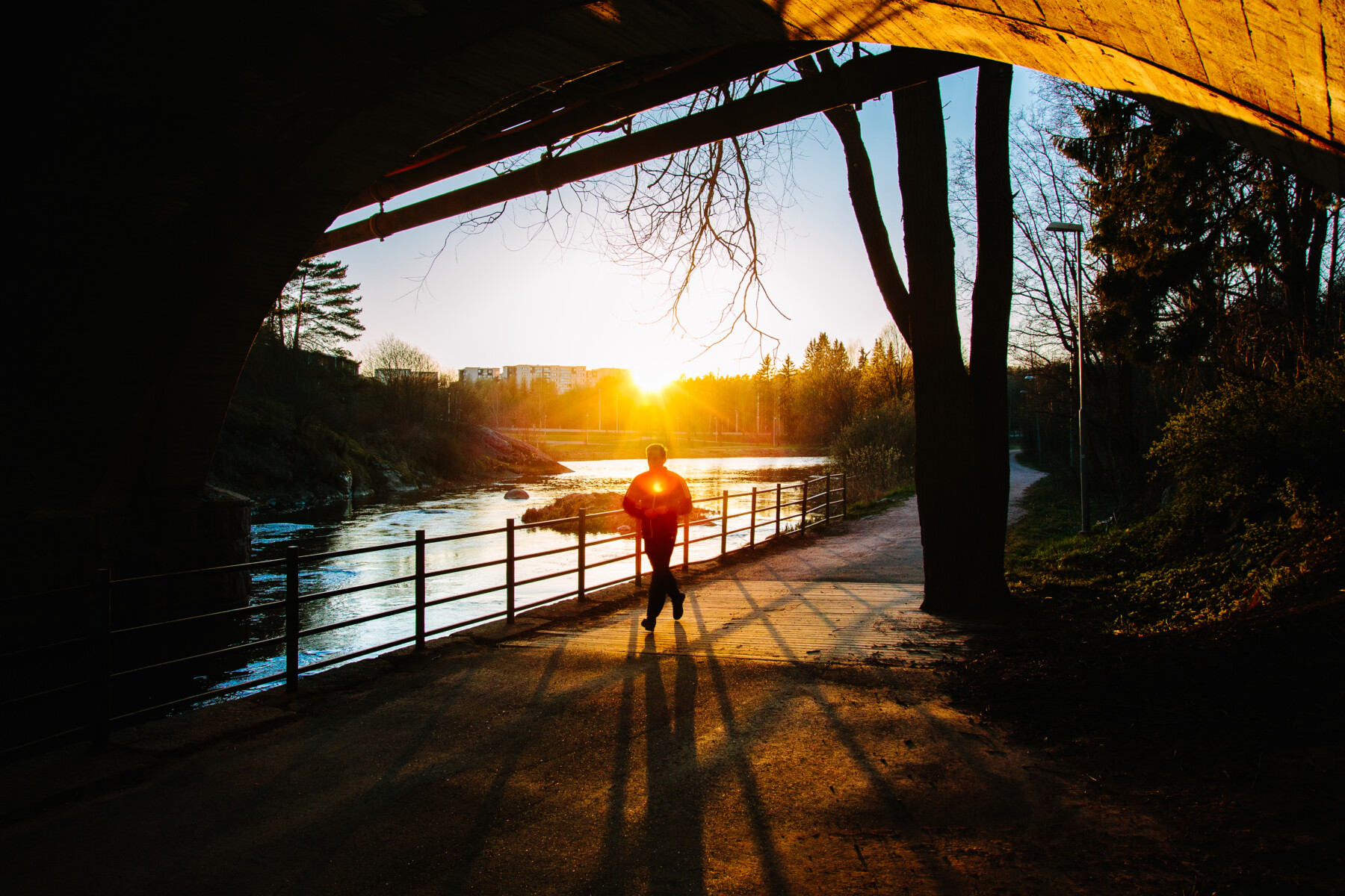 A person jogs along a riverside path beneath a bridge at sunset, their silhouette casting a long shadow across the ground.