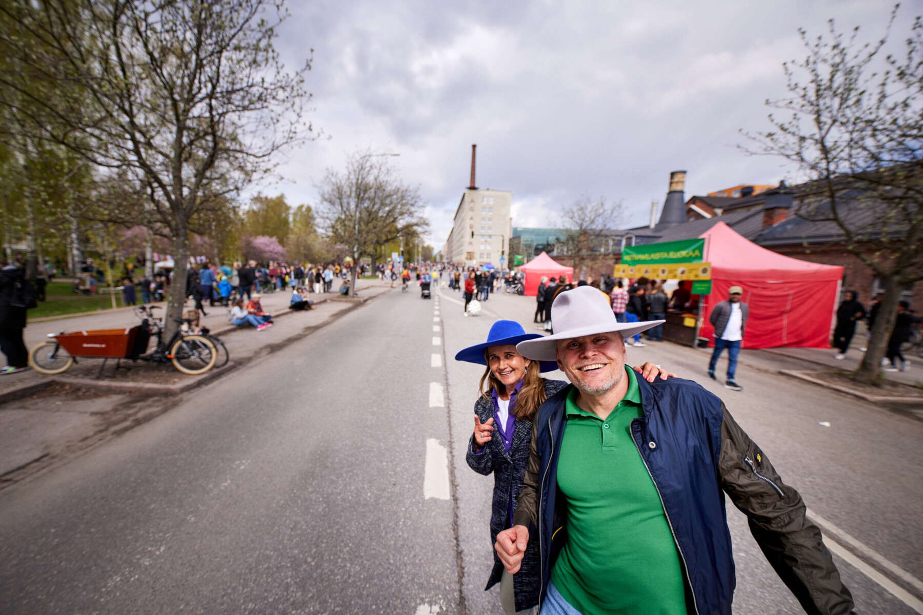 Two people in wide-brimmed hats smile at the camera during a street festival in Helsinki, with crowds and stalls along the road.