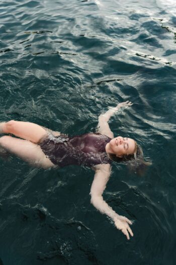 A woman floats on her back in deep, rippling water, wearing a one-piece swimsuit, with her arms outstretched and eyes closed.