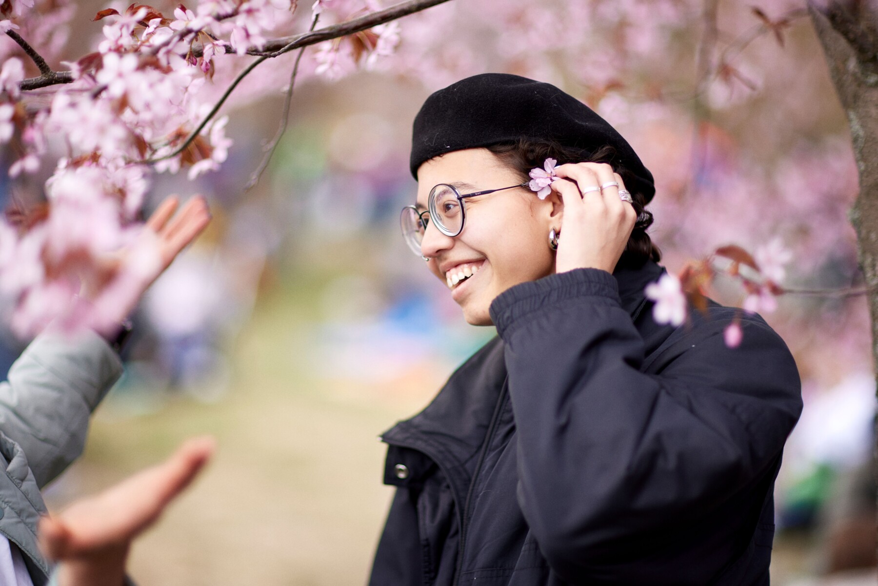 A smiling person wearing glasses and a black beret stands beneath pink cherry blossoms in Roihuvuori Cherry Park, Helsinki, Finland.