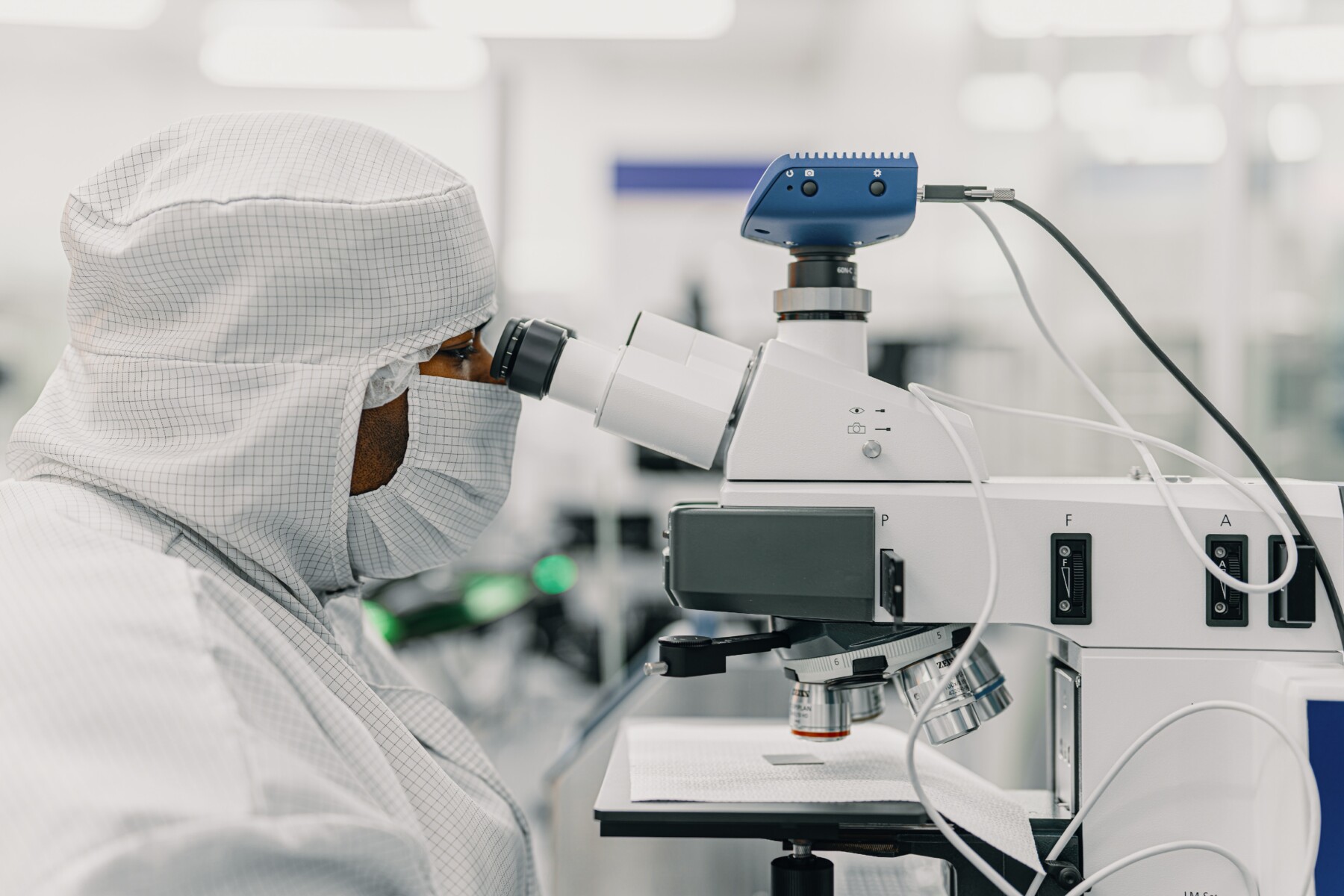 Une personne qui porte une combinaison de protection et un masque regarde dans un microscope à haute puissance, dans un laboratoire.