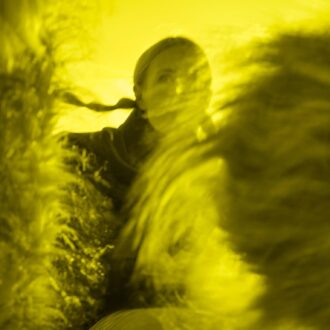 A woman moves amid blurred, windswept forms in a yellow-toned image, her face partially visible.
