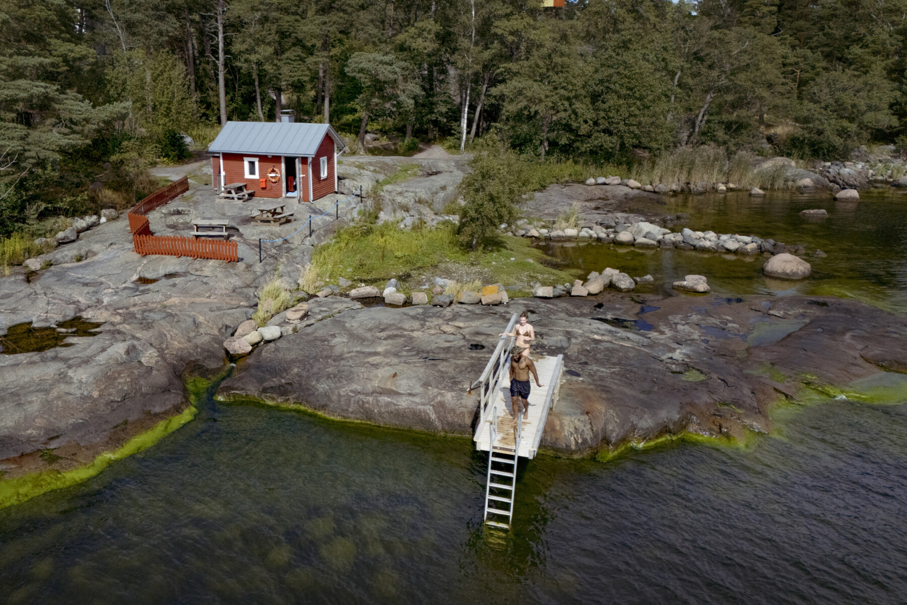 Two people stand on a small dock by a red cottage in Finland, preparing to swim along a rocky shore surrounded by forest.