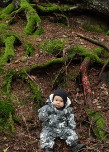 Um bebê agasalhado está sentado no chão da floresta, cercado por raízes de árvores cobertas de musgo e por folhas caídas.