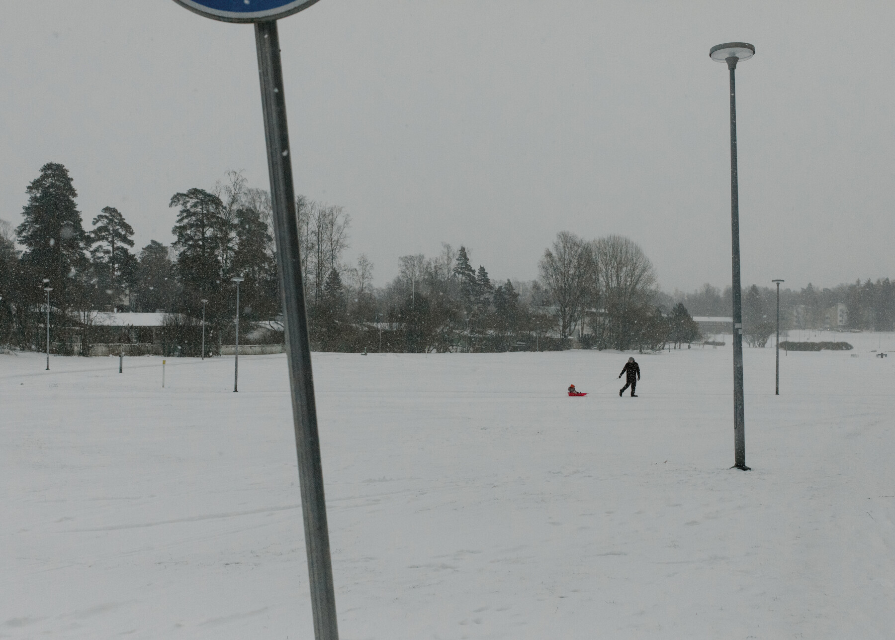 Uma pessoa puxa um trenó vermelho por um campo coberto de neve durante uma forte nevasca, com postes de luz altos e árvores ao fundo.
