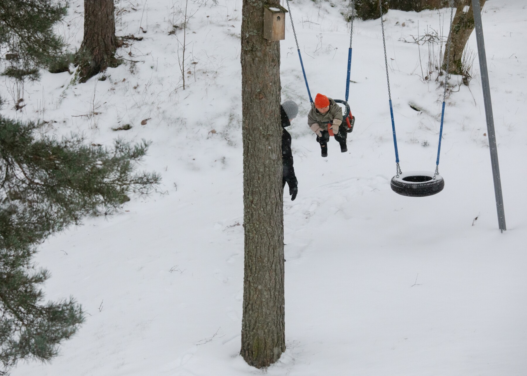 Uma criança com um chapéu laranja senta-se em um balanço em uma floresta nevada enquanto outra pessoa está por perto, parcialmente escondida atrás de uma árvore.