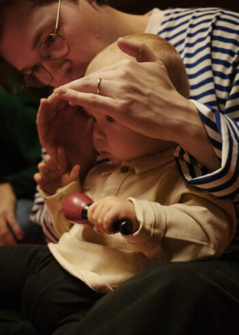 Um adulto de óculos protege delicadamente os olhos de uma criança pequena com as mãos enquanto ela segura um brinquedo pequeno.