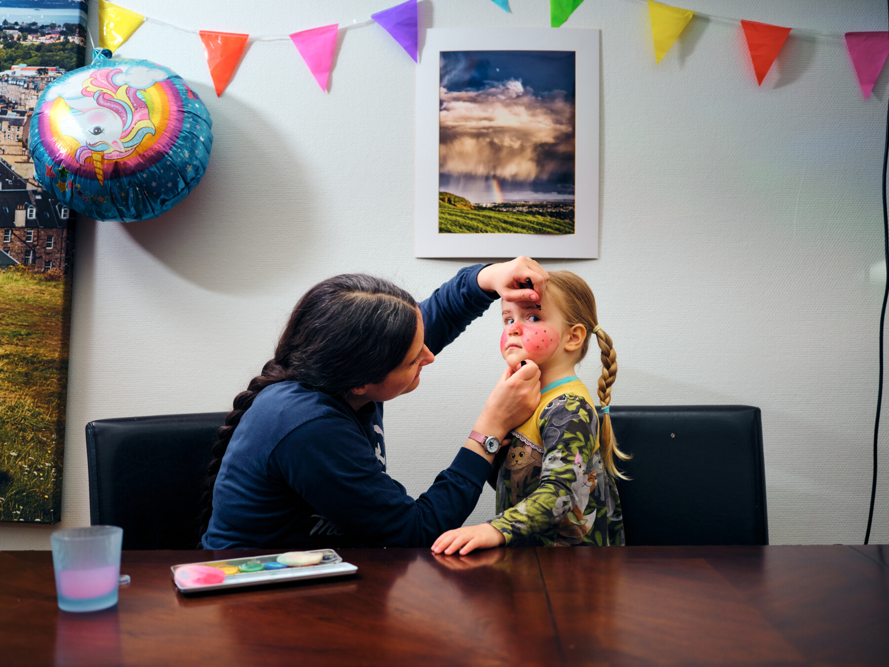 At a dining table, an adult carefully adds makeup to a child’s face as part of Easter preparations in Finland, with festive decorations around the room.