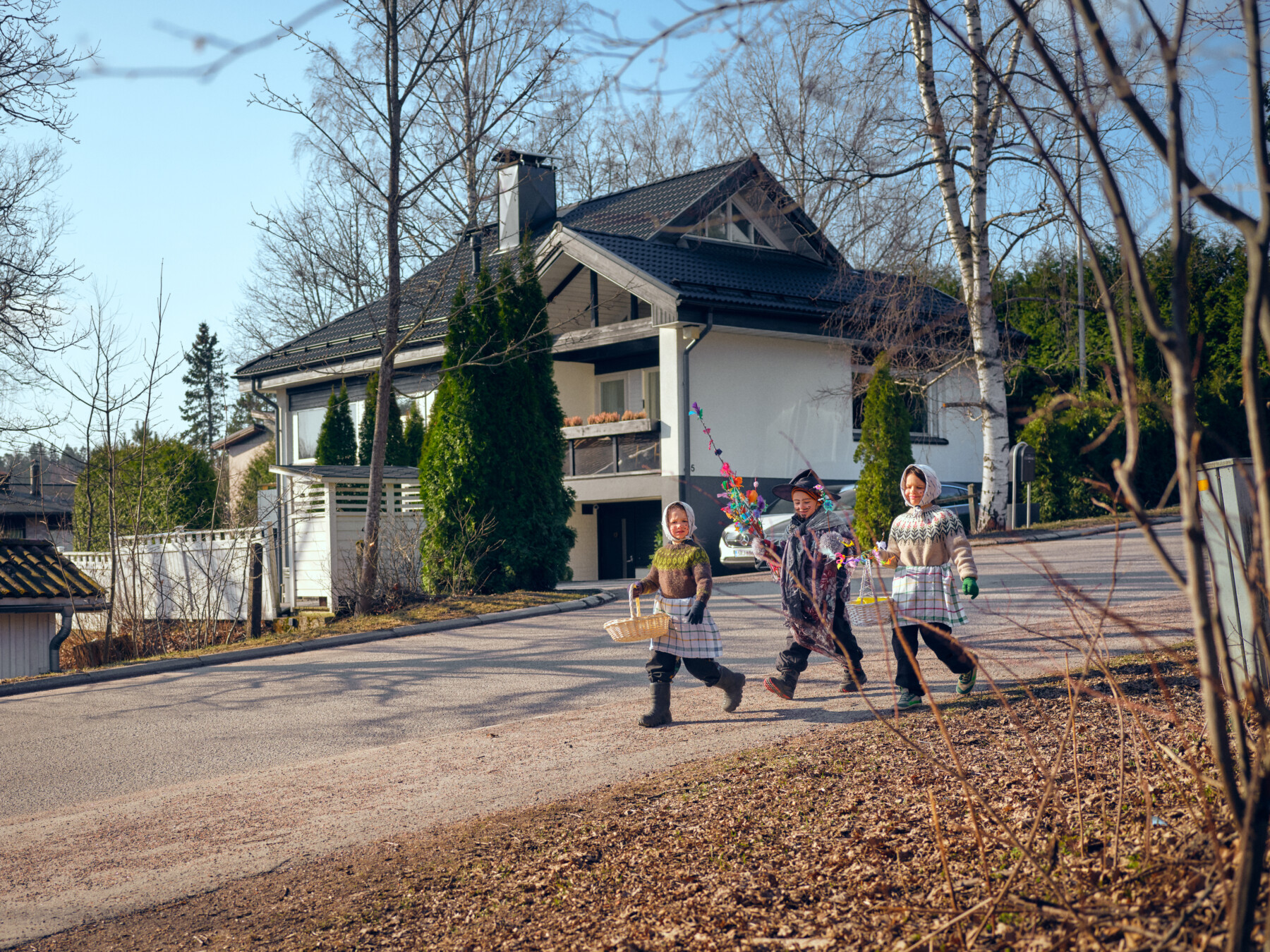 Three children in Easter witch costumes walk along a residential road carrying baskets and decorated willow branches.