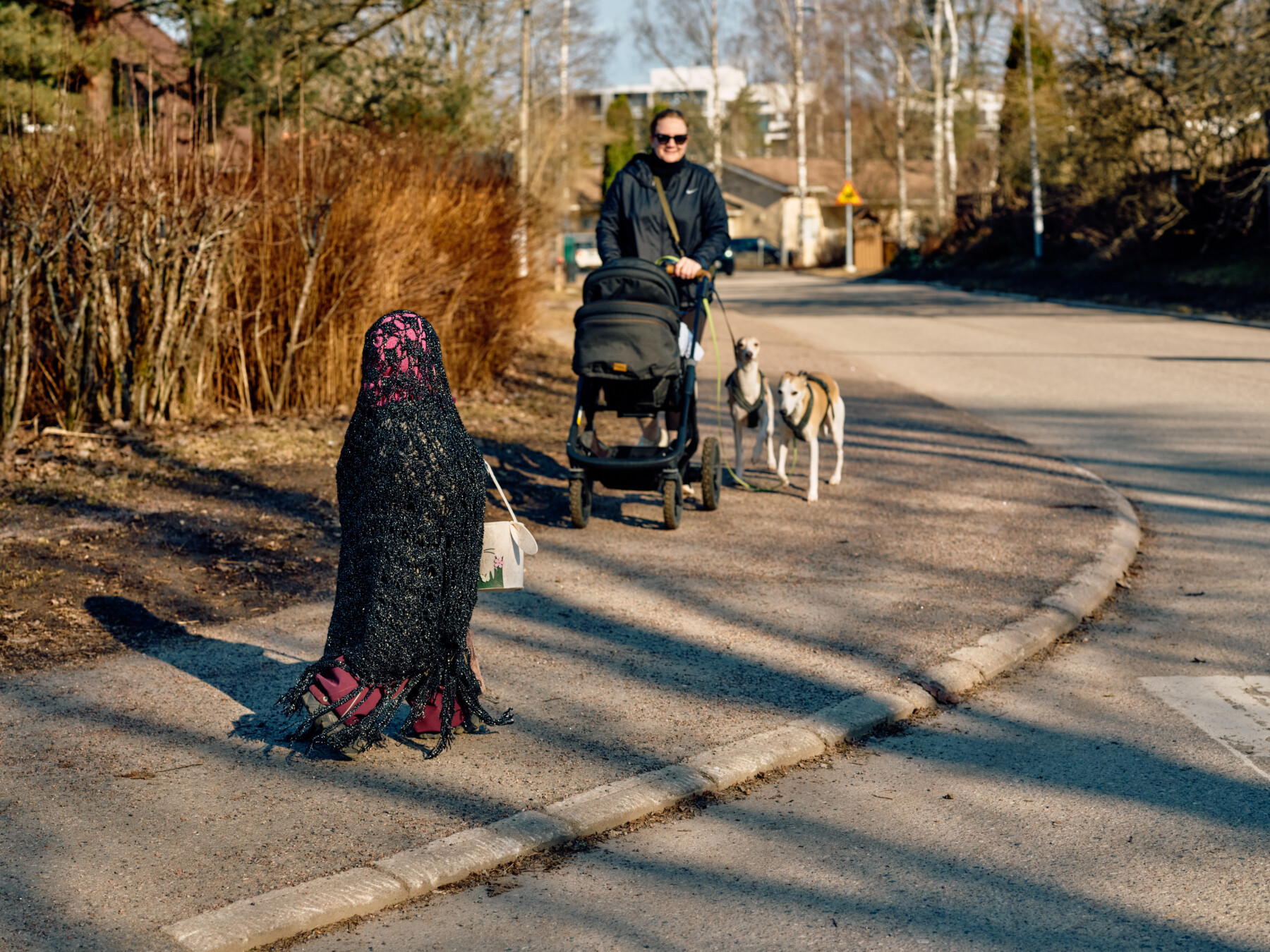 On a residential street in Finland, a child in an Easter witch outfit walks along the pavement as a neighbour with a pram and two dogs approaches.