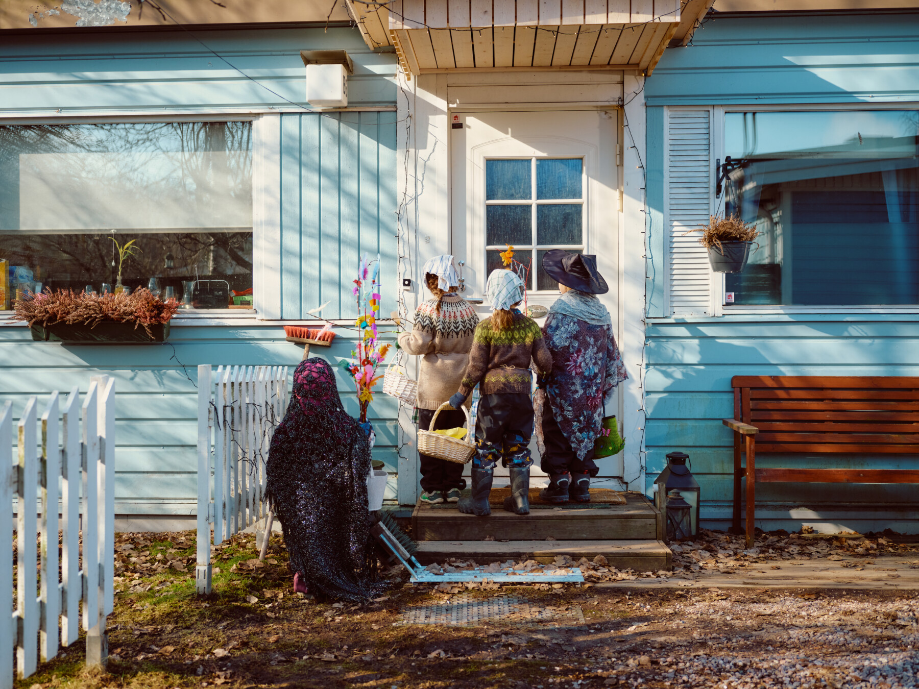 Children in Finland dressed as Easter witches stand at a front door holding decorated willow branches and baskets for sweets.