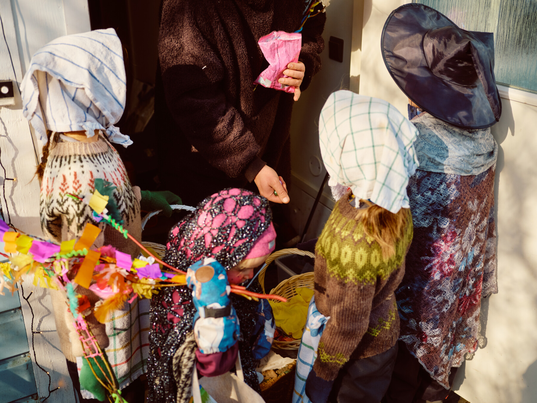 An adult gives treats to children dressed as witches at a front door, their baskets and colourful willow branches held nearby.