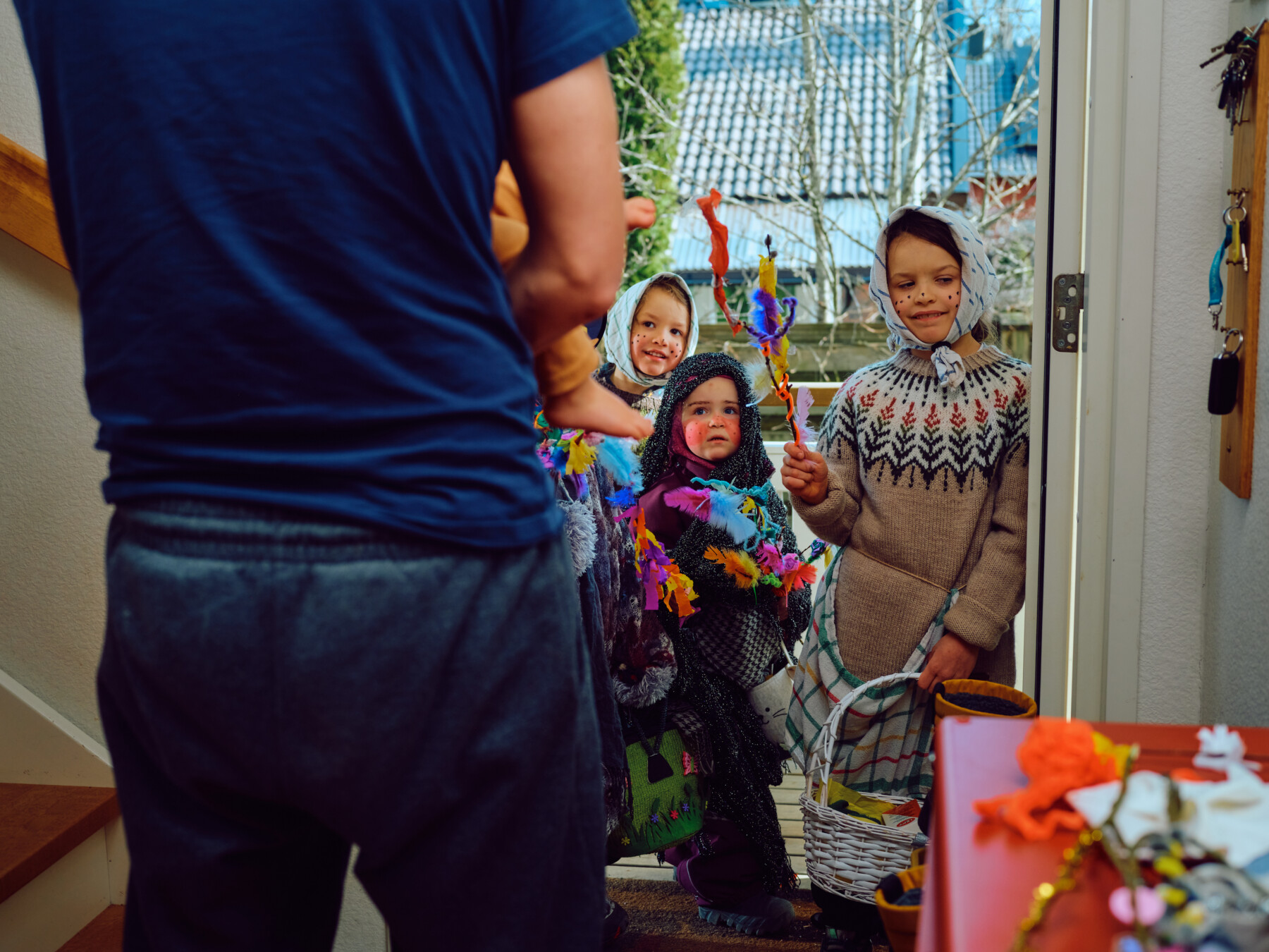 Children in Easter witch costumes stand at a doorway holding decorated willow branches while an adult answers from inside.