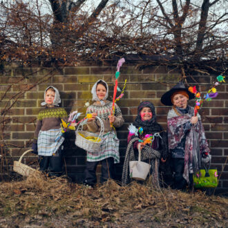 Four children dressed in Easter witch costumes stand in front of a brick wall in Finland, holding decorated willow branches and baskets.