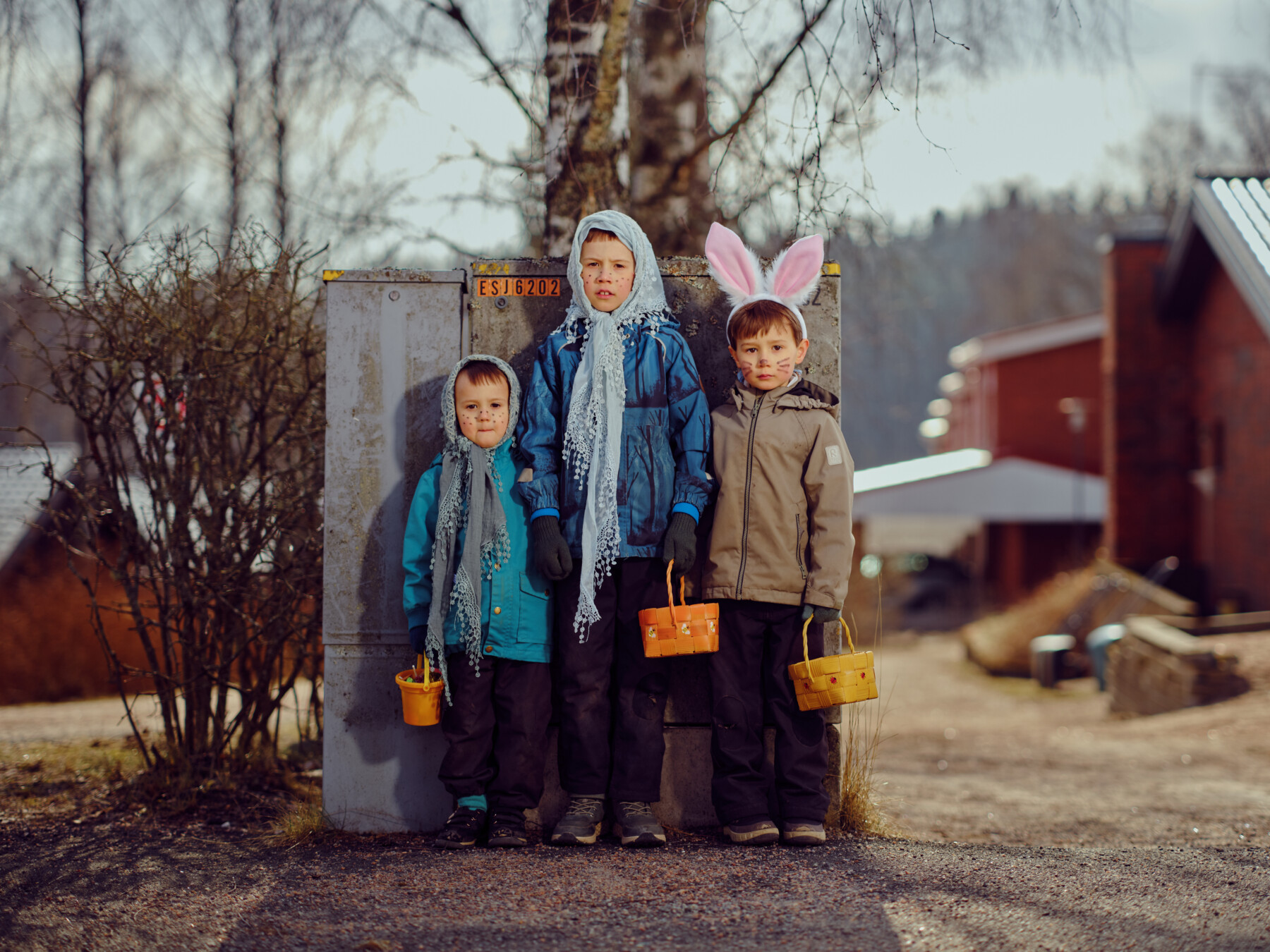 Children dressed for Finnish Easter traditions pose beside a utility box, carrying baskets and wearing headscarves and bunny ears.
