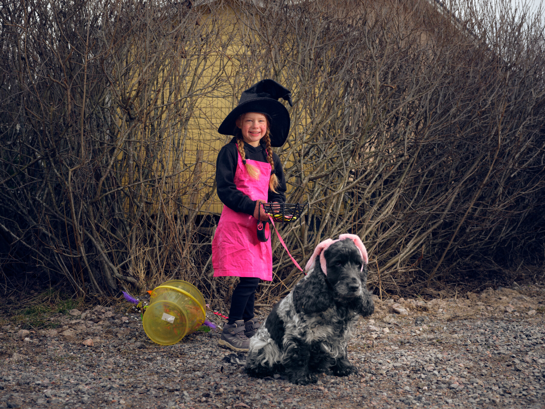 A child dressed as a witch in a pink outfit and black hat stands outdoors in Finland holding a basket and leash beside a dog wearing bunny ears.