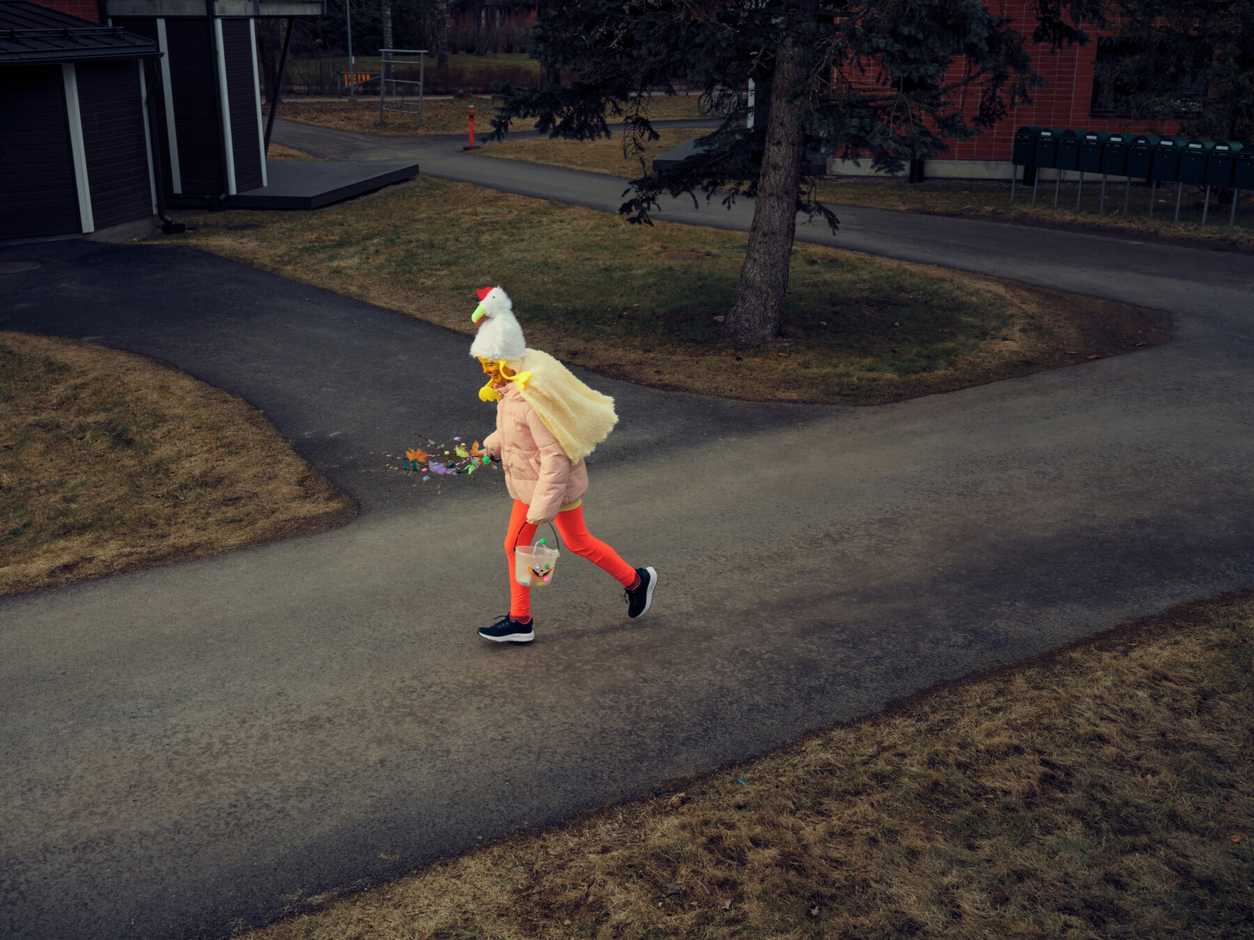 A costumed child with a hat and cape carries willow twigs and sweets while hurrying through a neighbourhood during Finnish Easter traditions.