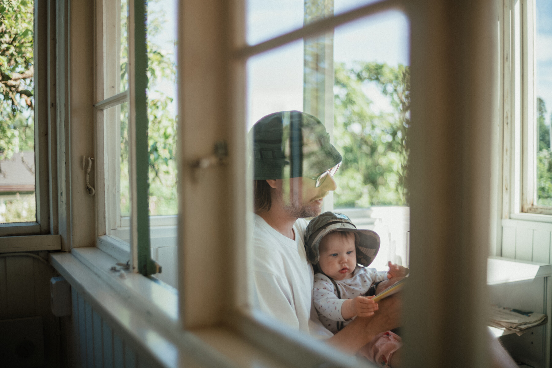 A baby wearing a sun hat sits on an adult’s lap inside a bright wooden cottage with open windows and trees visible beyond.