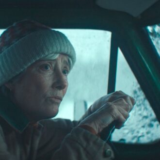 La actriz Emma Thompson con un gorro de invierno en una fotografía de la película Muerte en invierno al volante de un coche con un paisaje forestal nevado visible a través de la ventanilla.