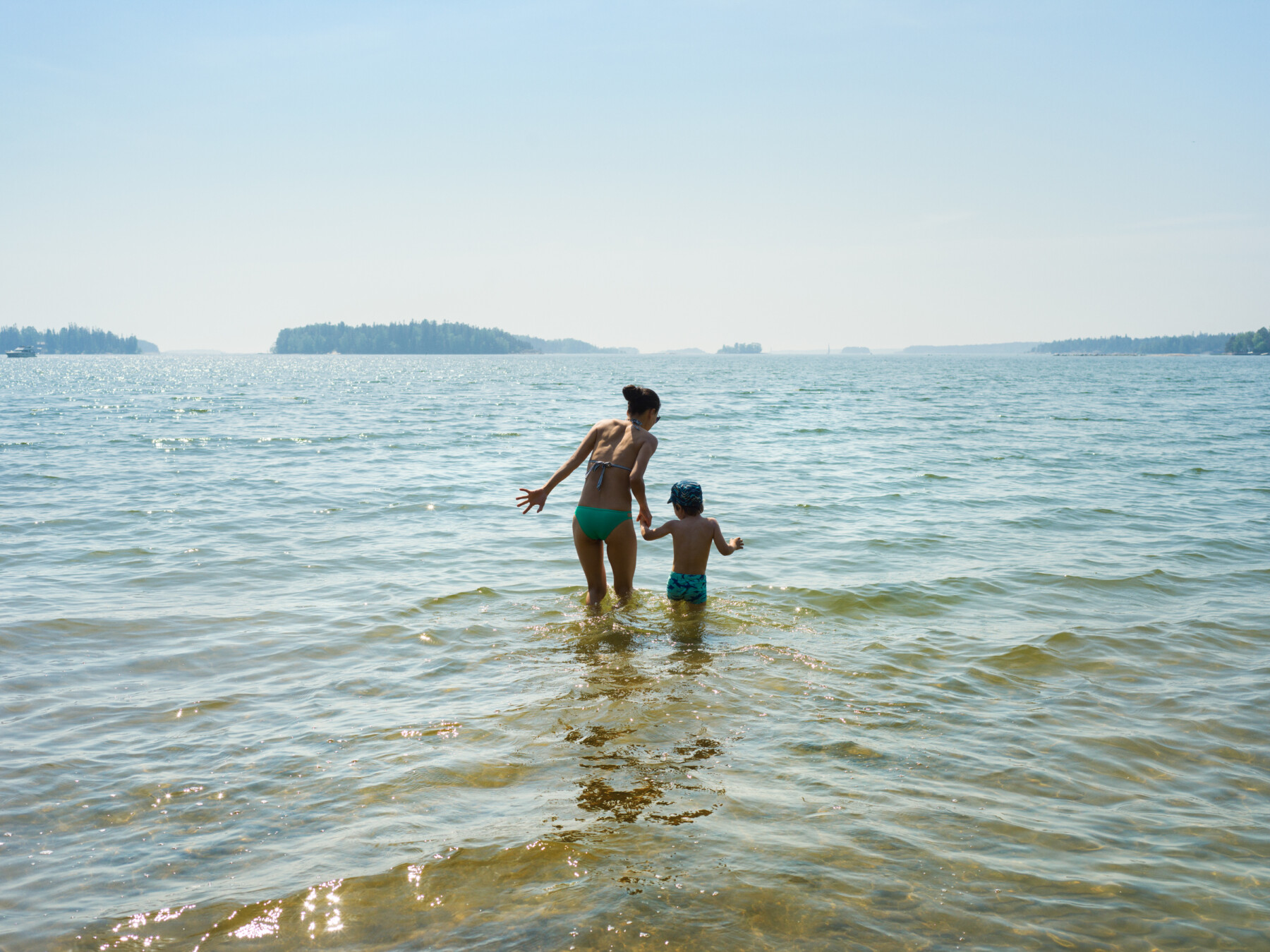 An adult and child wade through shallow water near the shore of a lake, with small, forested islands visible in the distance.