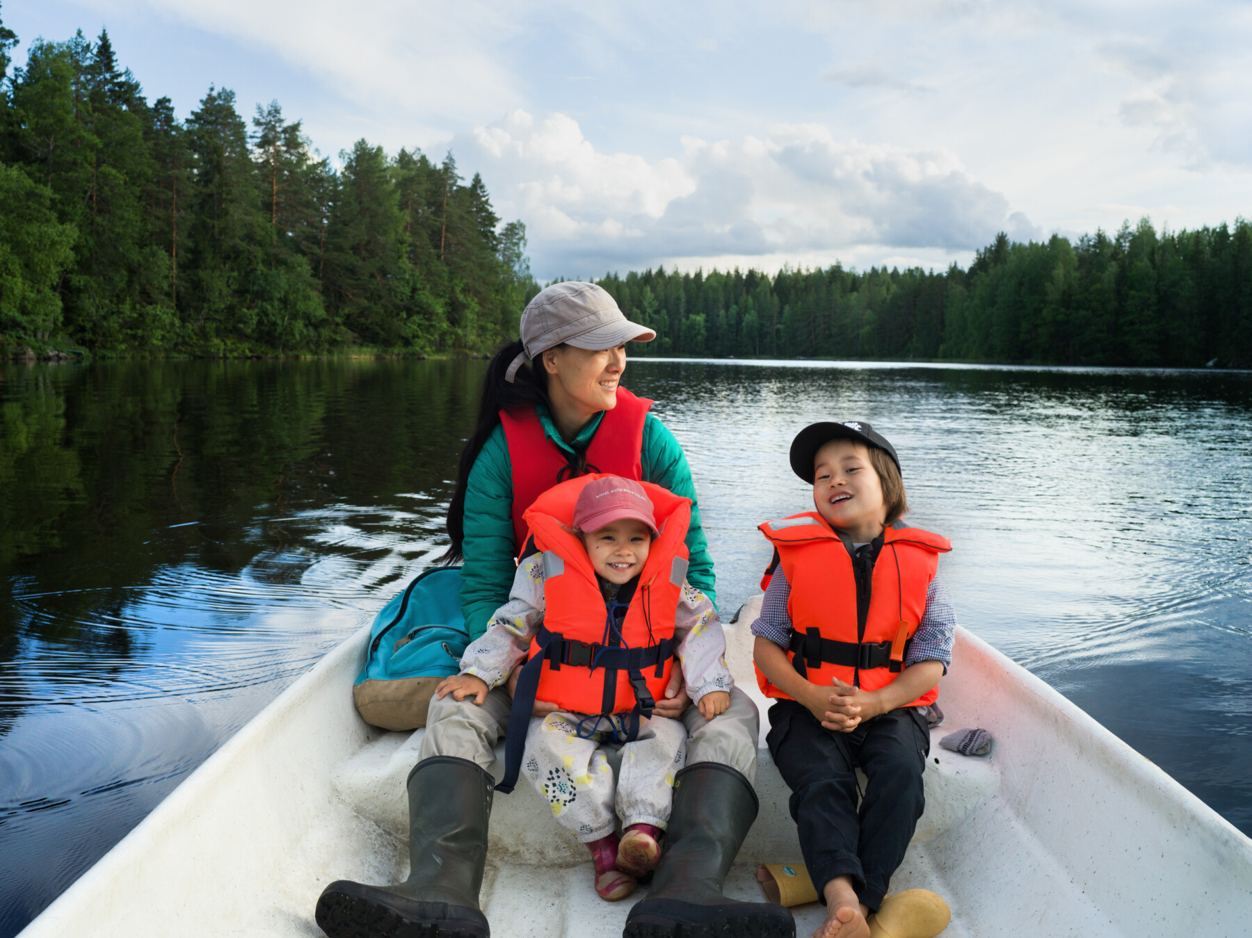 Two young children in orange life jackets ride in a rowing boat with an adult on a peaceful lake surrounded by dense woodland and cloudy skies.
