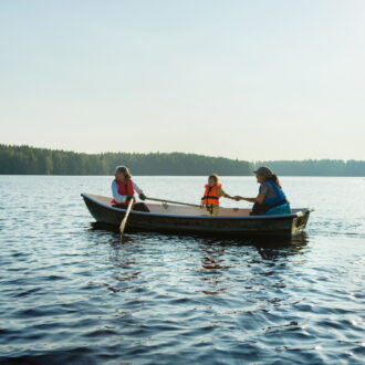 Two adults and a child wearing life jackets row a small boat across a calm Finnish lake surrounded by forest in the evening light.