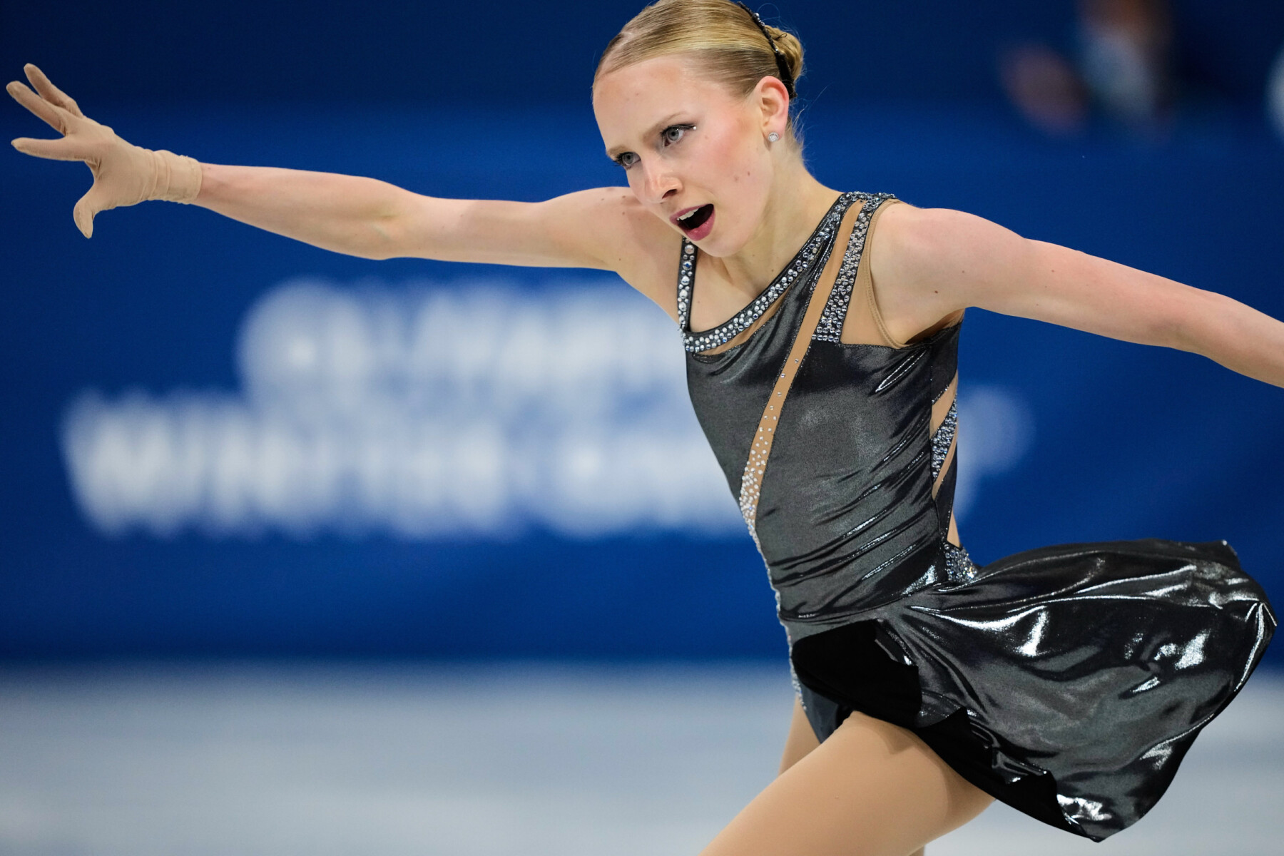 Iida Karhunen skates in a black costume with arms extended as her skirt ripples in motion behind her.