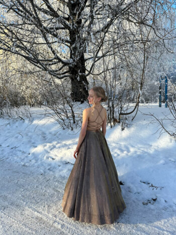 Finnish figure skater Iida Karhunen wears a floor-length gown and stands on a snowy path beneath frost-covered trees, looking over her shoulder.