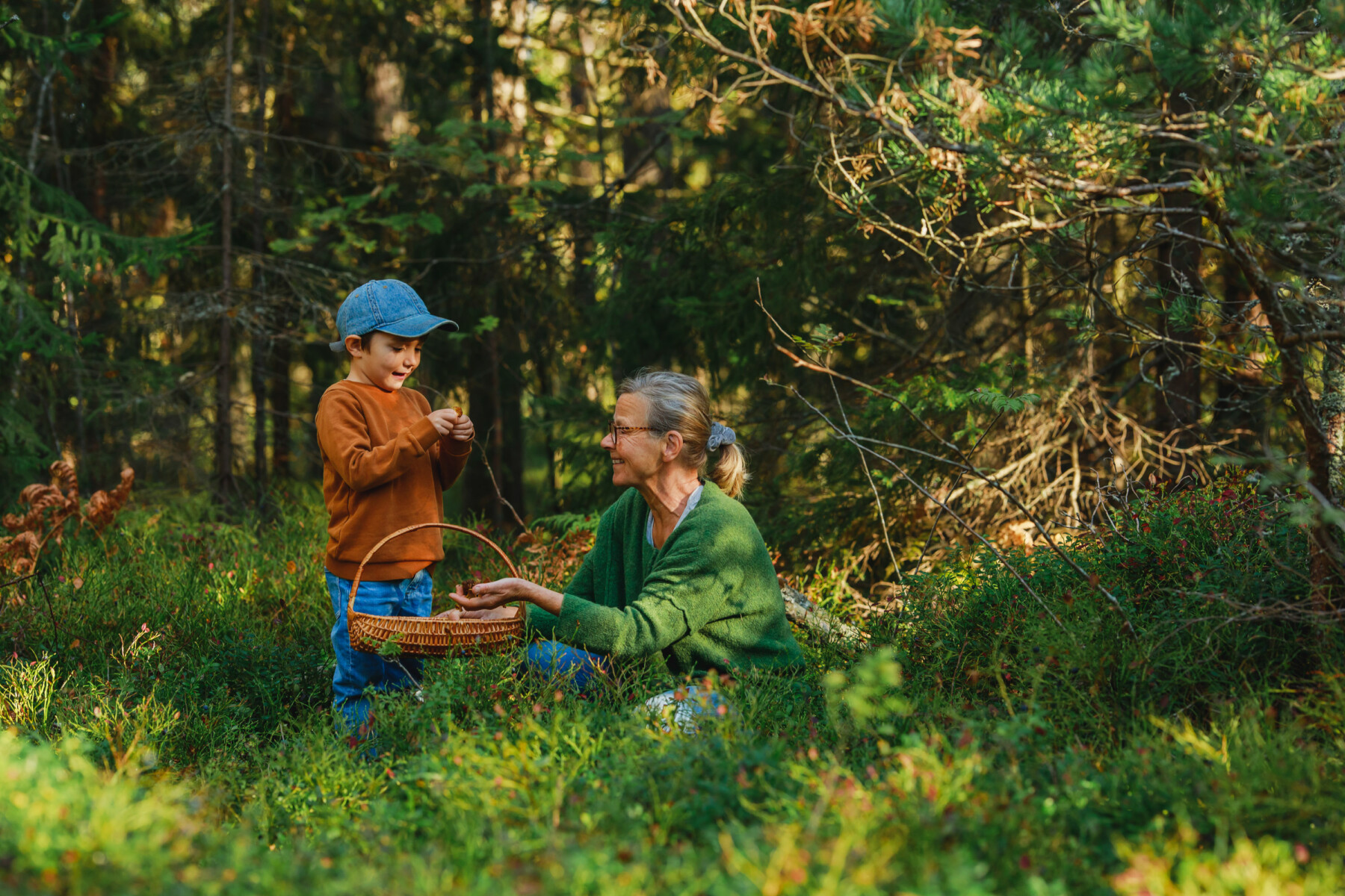 An adult and child gather berries in a sunlit forest clearing, with a woven basket resting between them.