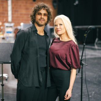 Pete Parkkonen avec ses cheveux bouclés et Linda Lampenius avec sa longue chevelure blonde se tiennent côte à côte dans des tenues sombres avec l’équipement scénique en arrière-plan.