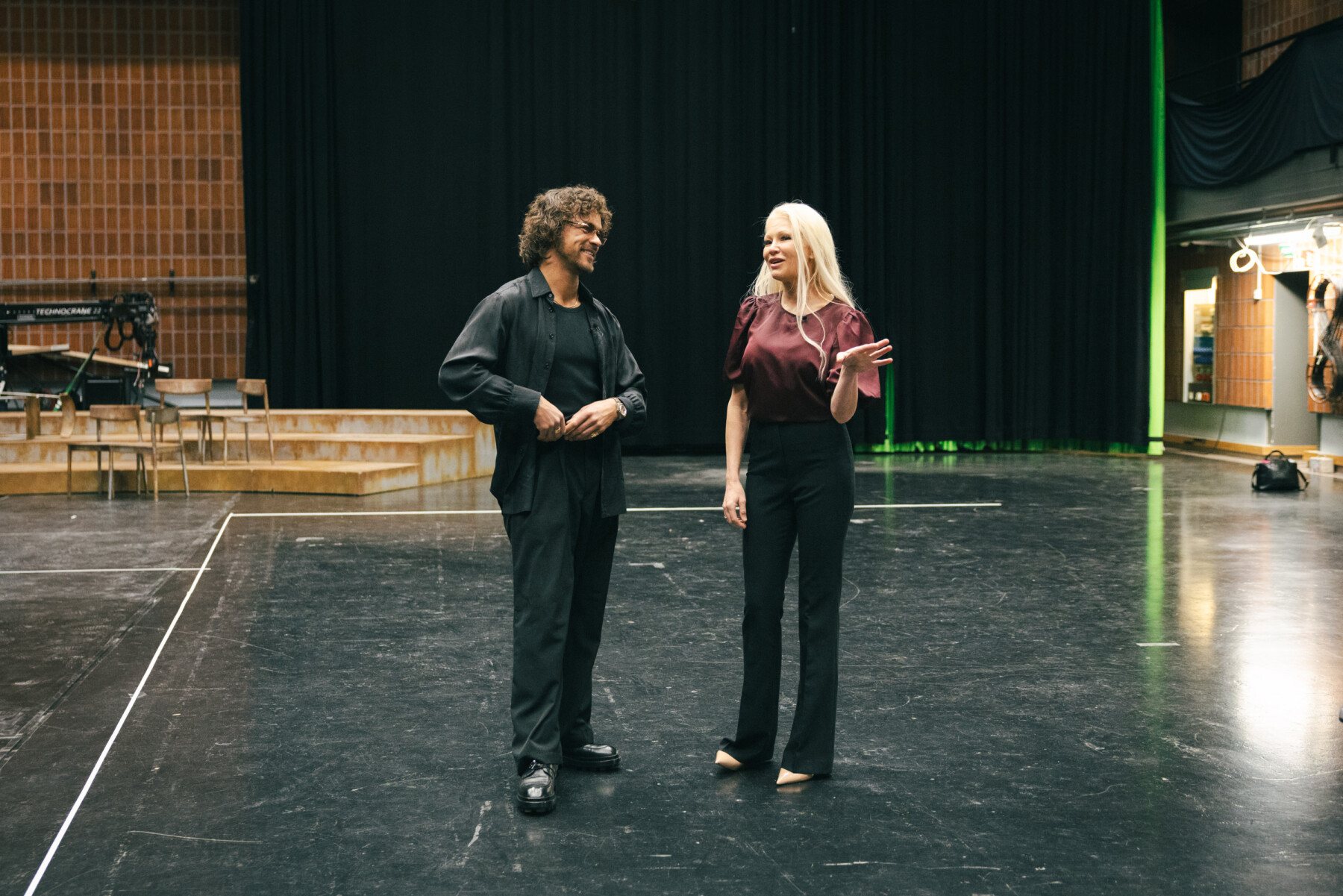 Linda Lampenius et Pete Parkkonen se font face sur une scène vide et discutent avec des fauteuils de théâtre et des équipements en arrière-plan.