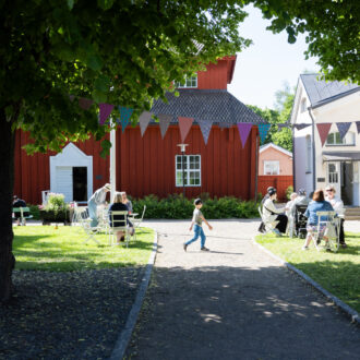 Ein Kind läuft einen Kiesweg entlang, während sich Menschen an Tischen im Freien in einem grünen Innenhof im Museumsviertel von Neristan versammeln.