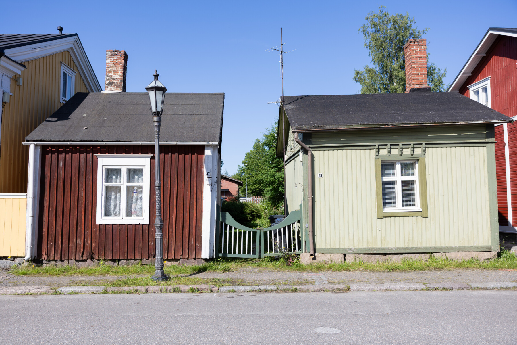 Two small wooden houses in Neristan stand side by side, separated by a narrow gate and a traditional street lamp.