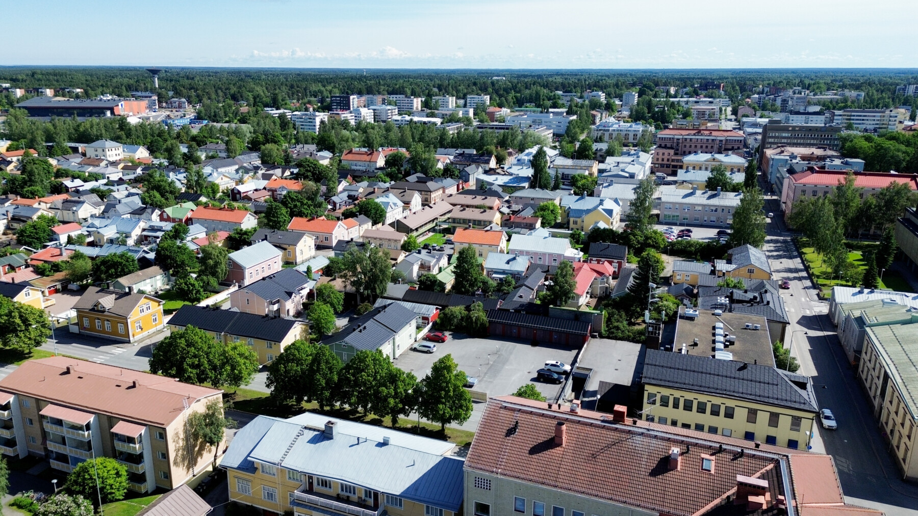 An aerial view of Neristan shows a grid of pastel wooden houses, tree-lined streets and low historic buildings stretching towards the modern city beyond.