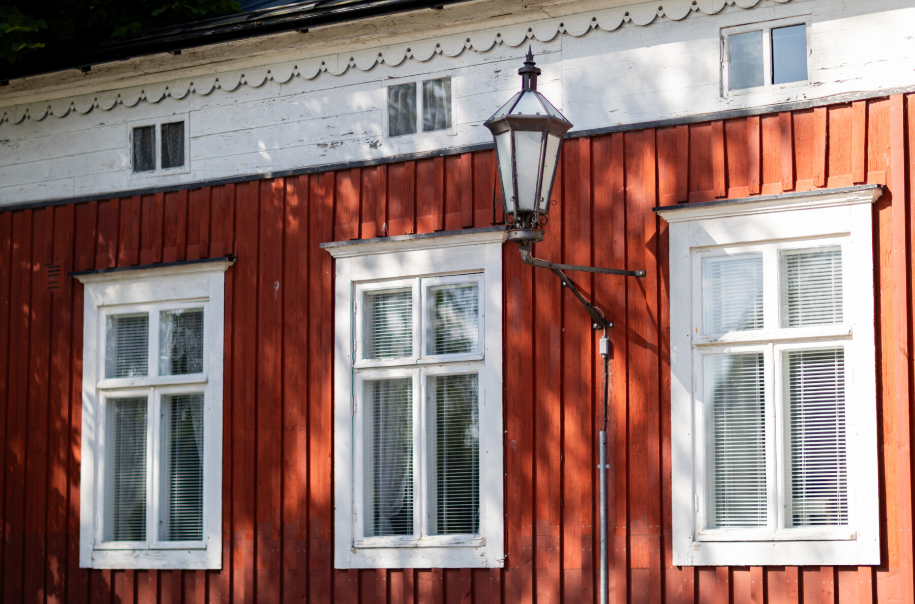 A red wooden house in Neristan features white-framed windows and an old-fashioned wall-mounted lantern.