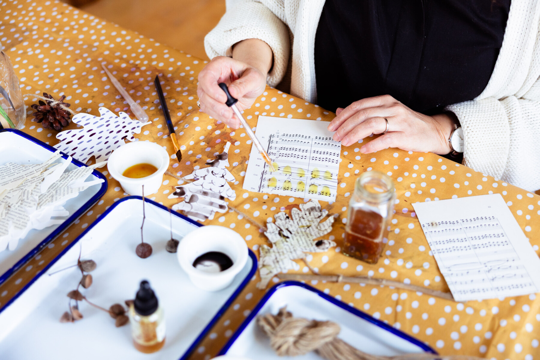 A close-up of hands painting with natural ink on old sheet music, with small trays of natural materials and ink bottles arranged on a yellow polka-dot tablecloth.