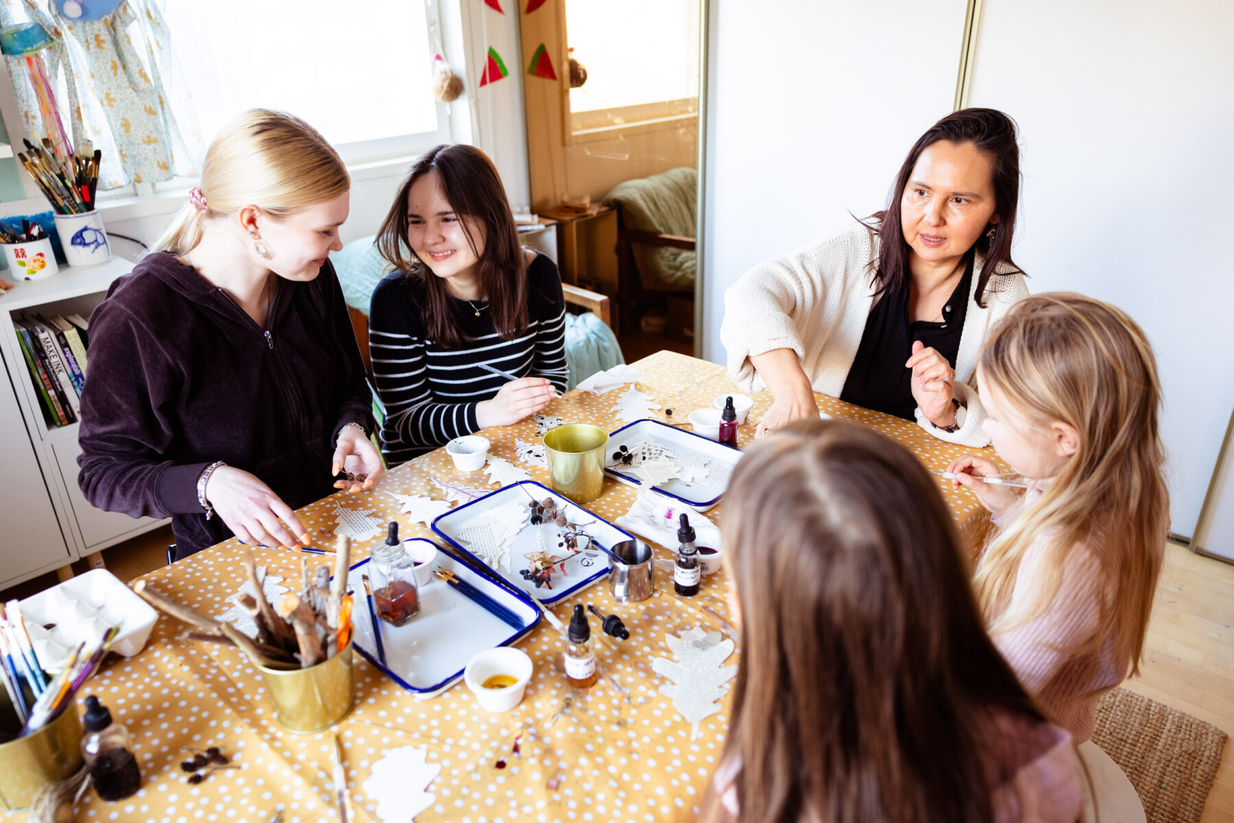 A woman, Rushan Nasretdin, sits at a table with two children and two teenagers during a natural ink workshop, everyone engaged with materials spread across a yellow tablecloth.