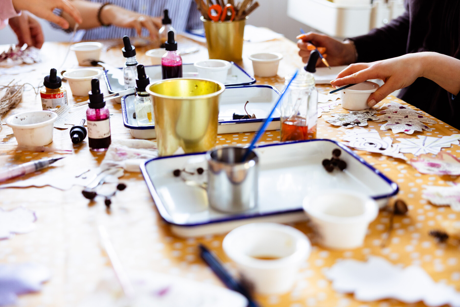 A close-up of a workshop table covered with ink bottles, small cups, paper cut-outs and trays of natural materials, with hands reaching in from the sides.