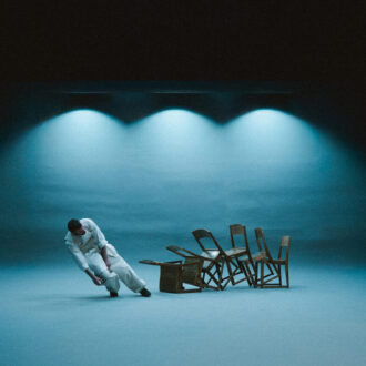 A performer in white leans at an angle on a minimalist stage as several wooden chairs appear to tilt and defy gravity under cool blue lighting.