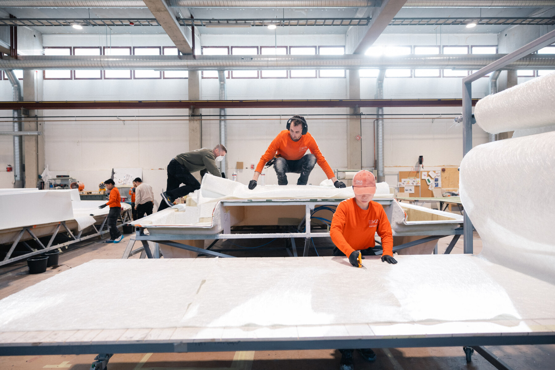 Arbeiter in orangefarbenen Hemden handhaben leichte Materialien bei der Herstellung von Bootskomponenten in einer großzügigen Halle bei Eka Composite.